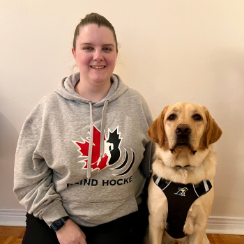 Laura and her CNIB Guide Dog, Toby. Indoors, Laura kneels on the ground next to Toby, a yellow lab in harness. Laura smiles and has her left arm around Toby. Laura is also wearing a Canadian Blind Hockey sweater. 