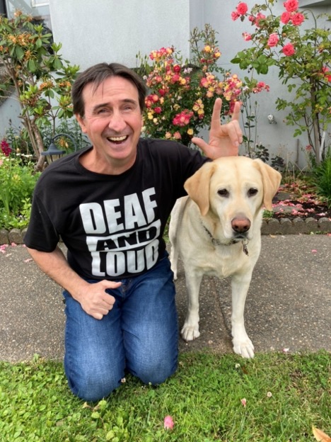 Eddy sits in front of a backyard garden with his guide dog. His left-hand hovers above the dog’s head as he signs “I love you” in ASL. The black t-shirt Eddy is wearing has a bold text design that reads “Deaf and loud!” 