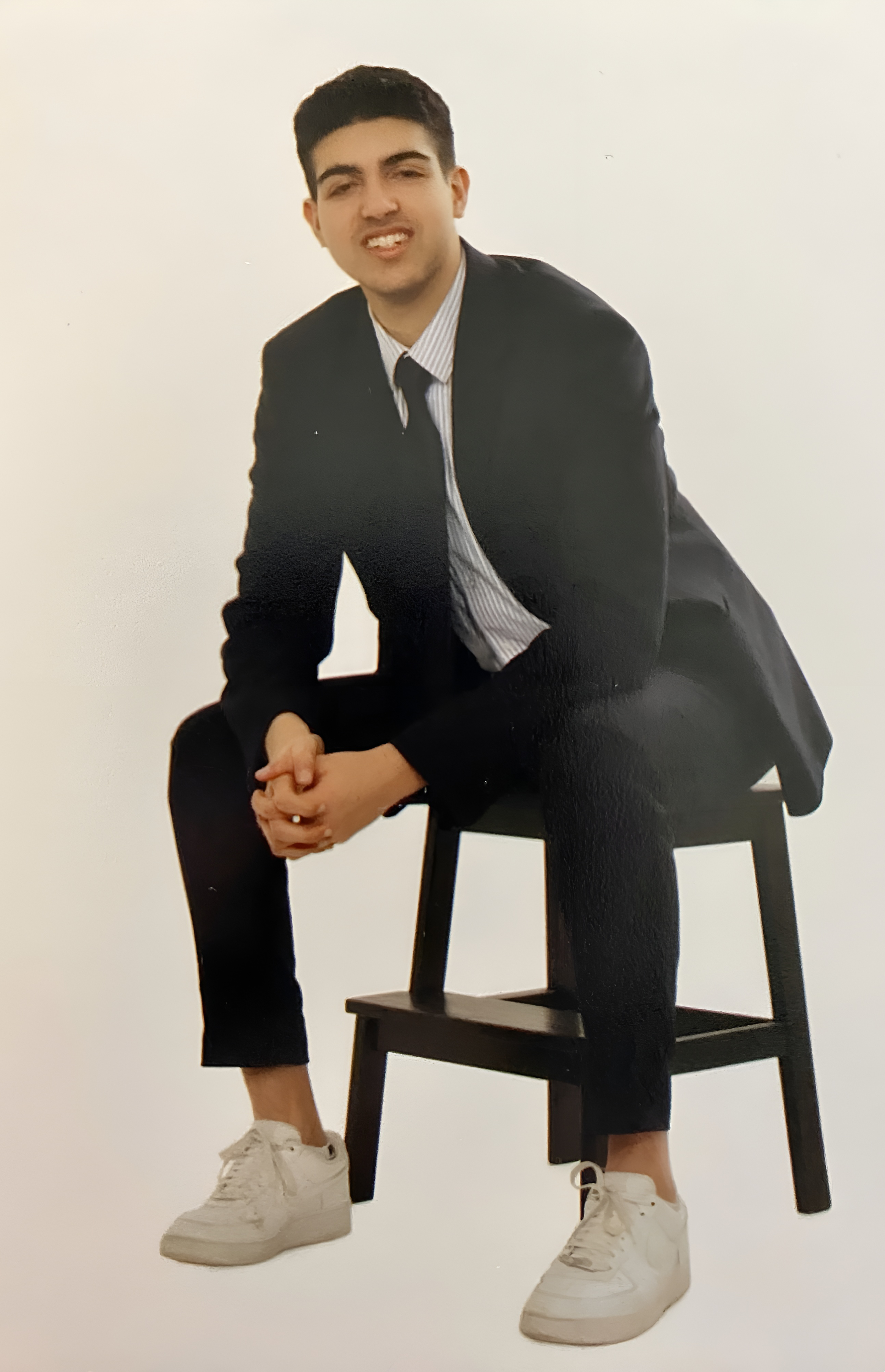 A professional photo of Aidan. He is wearing a black suit and tie with a pair of white sneakers and sits on a stool with his hands clasped in his lap.