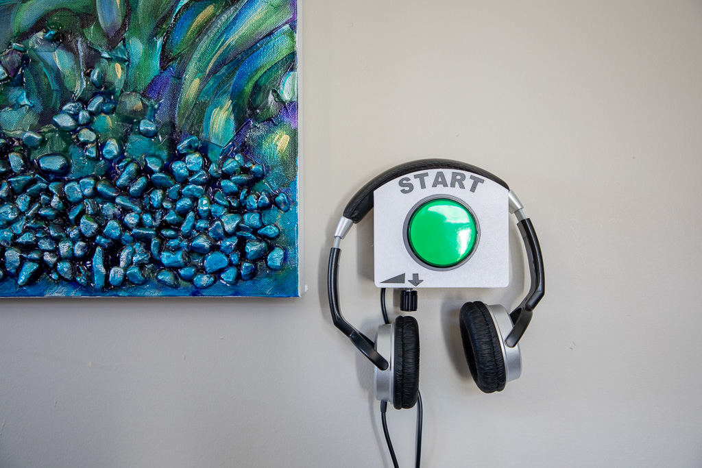 Une peinture abstraite en bleu et vert est accrochée au mur. À côté de la peinture, sur le mur, se trouve un appareil muni d’un gros bouton vert surmonté du mot « Start ». Une flèche pointe vers le bas vers un bouton. L’appareil est surmonté d’un casque d’écoute.