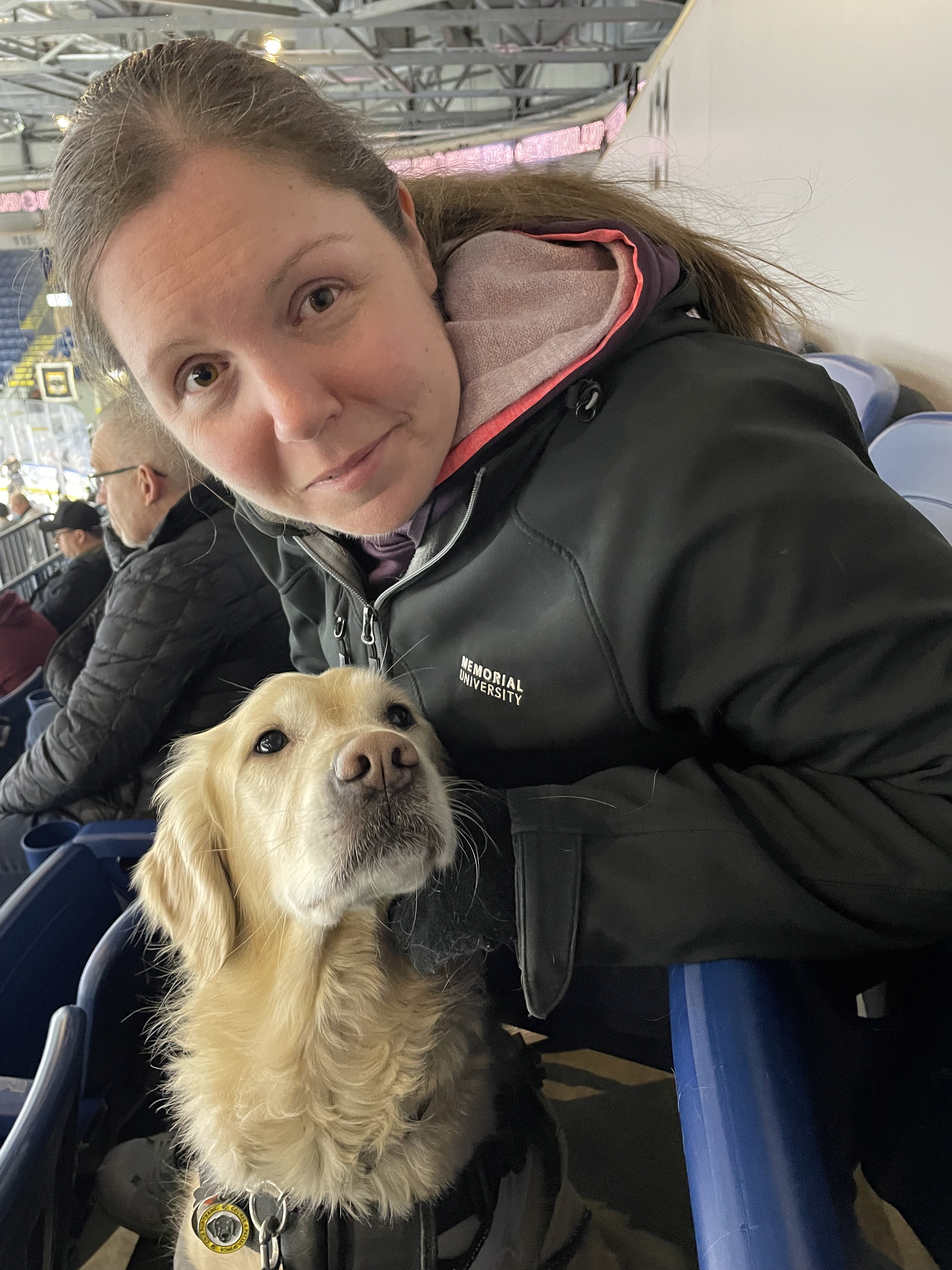 Kelly and her guide dog, Maple, sit in the stands at a hockey arena. 