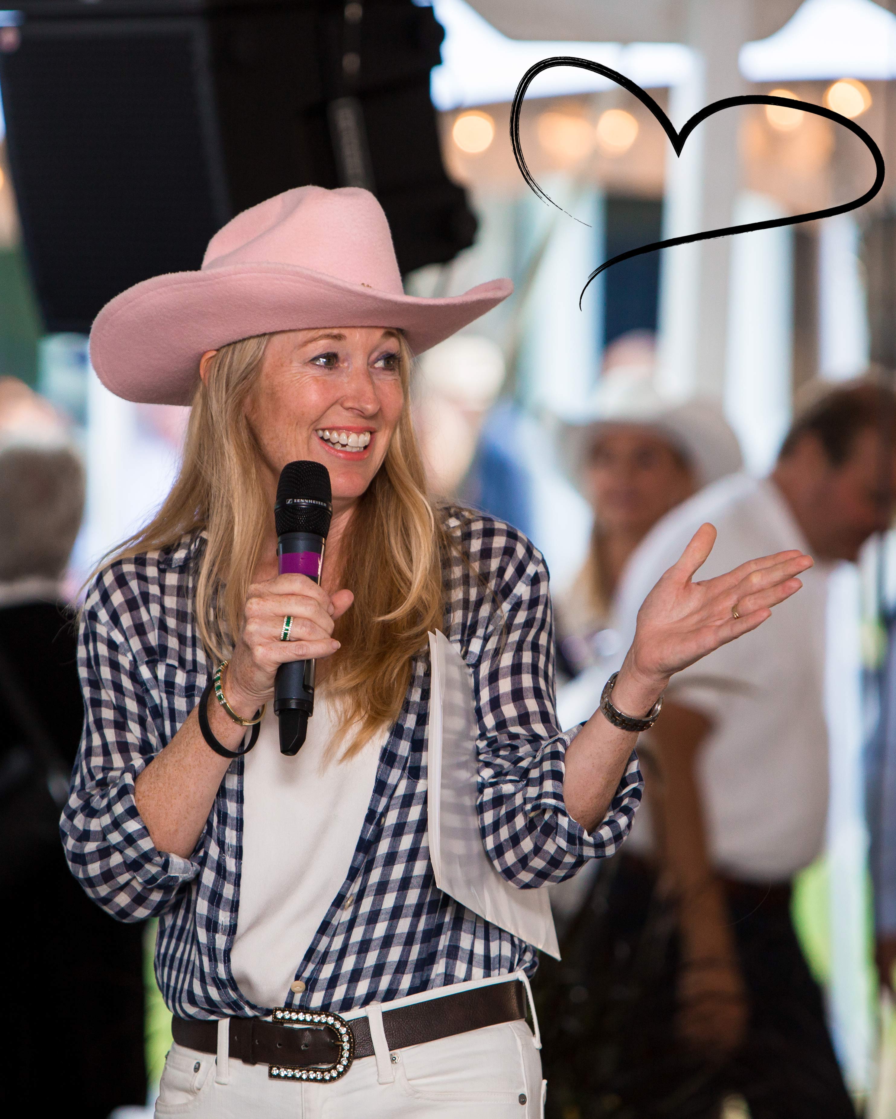 Nancy Simonot, portant un chapeau de cowgirl rose et une chemise à carreaux, un micro à la main, s'adresse à une foule. Un dessin en forme de cœur apparaît sur la table, dans le coin inférieur droit supérieur de l'image