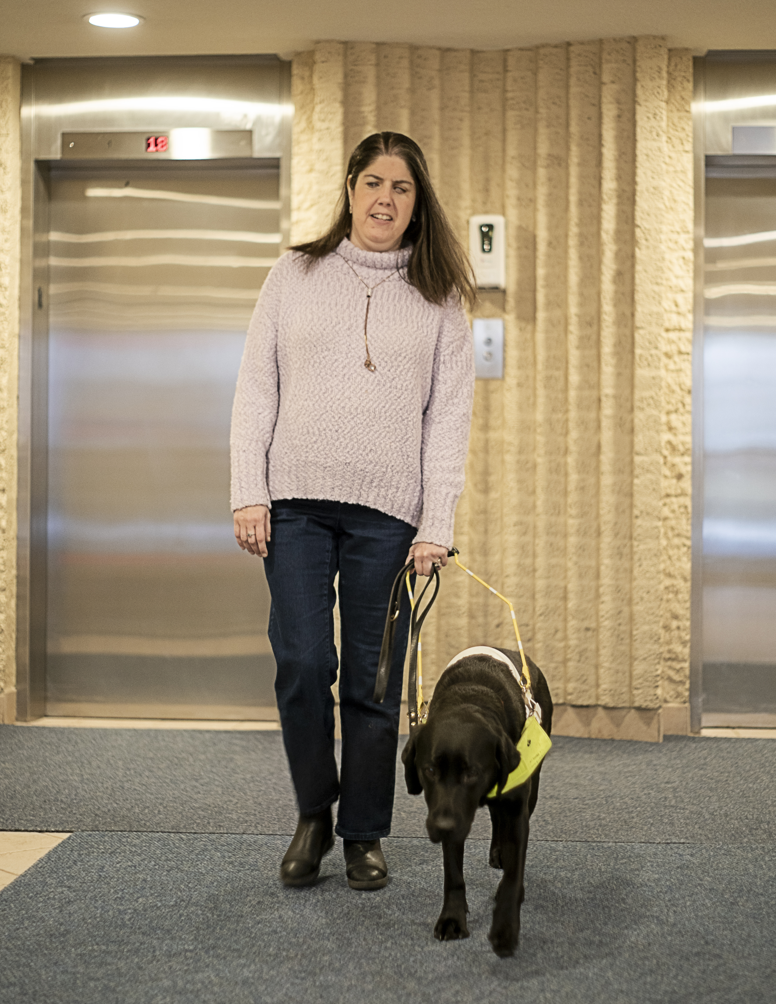 Karoline et son chien-guide noir, Raven, sortent du hall d’un immeuble. Derrière eux se trouve une rangée d’ascenseurs.