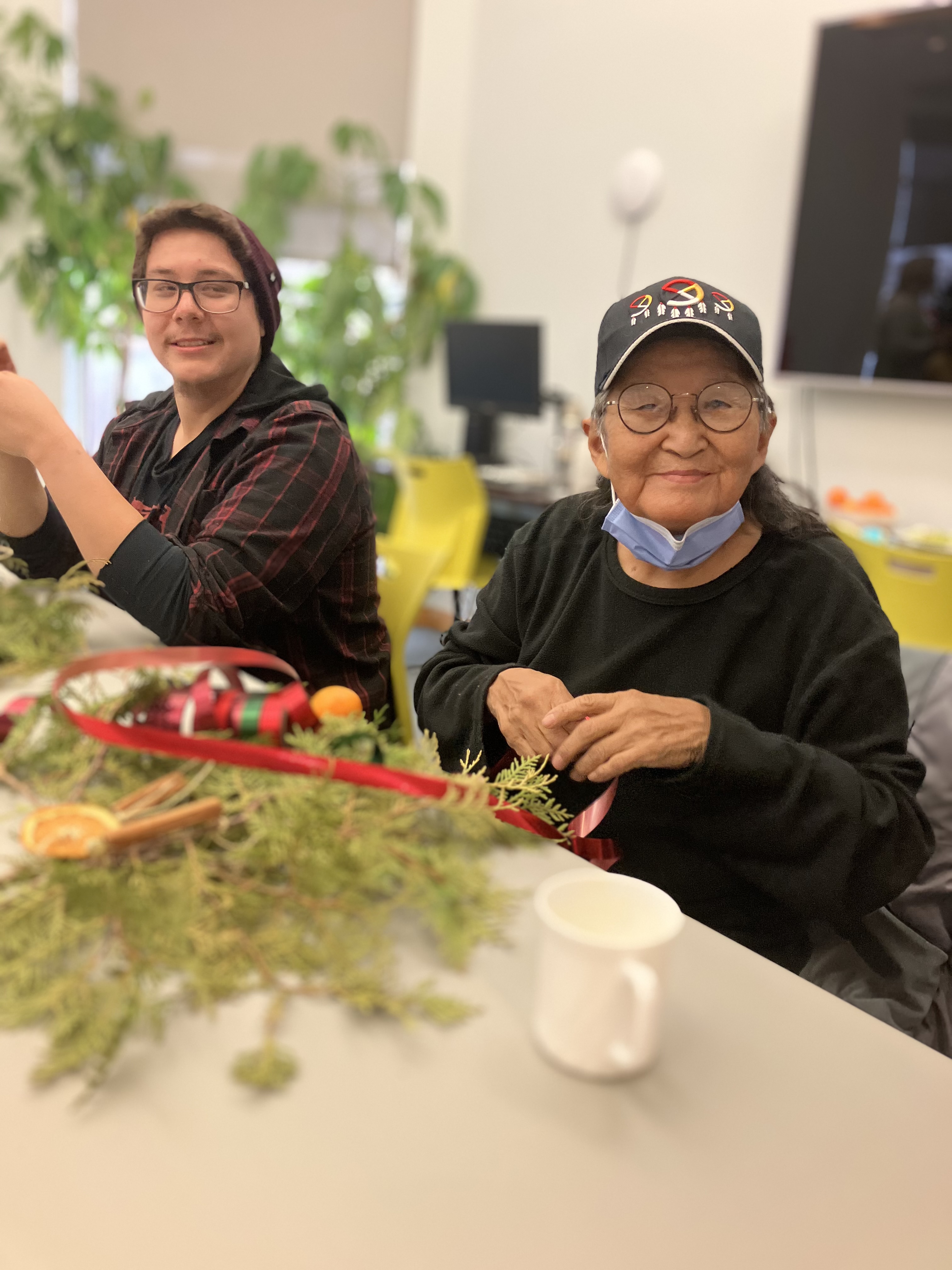 Two Indigenous participants sit at a table in CNIB Thunder Bay smiling. During a holiday program, they create decorations out of fresh cedar bows, dried orange slices, homemade gingerbread cookies, ribbons, bells, cinnamon sticks, and jute twine.