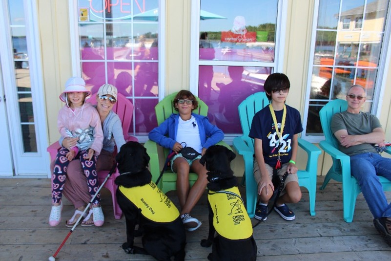 12 year-old Gabriel sits in a colourful Muskoka chair, relaxed and smiling, surrounded by other happy CNIB Lake Joe campers and families, and two CNIB buddy dogs in yellow vests.