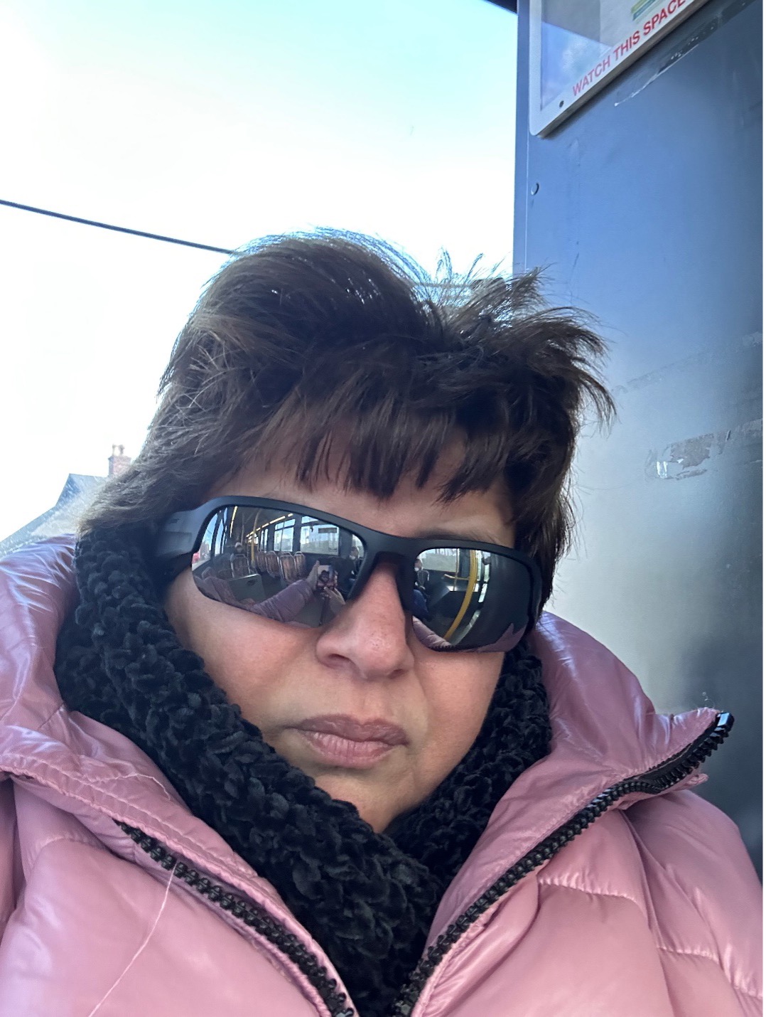 Marisa rides the bus. She’s wearing sunglasses, a pink winter coat and a black knitted scarf. The interior of a bus is reflected in her sunglasses.
