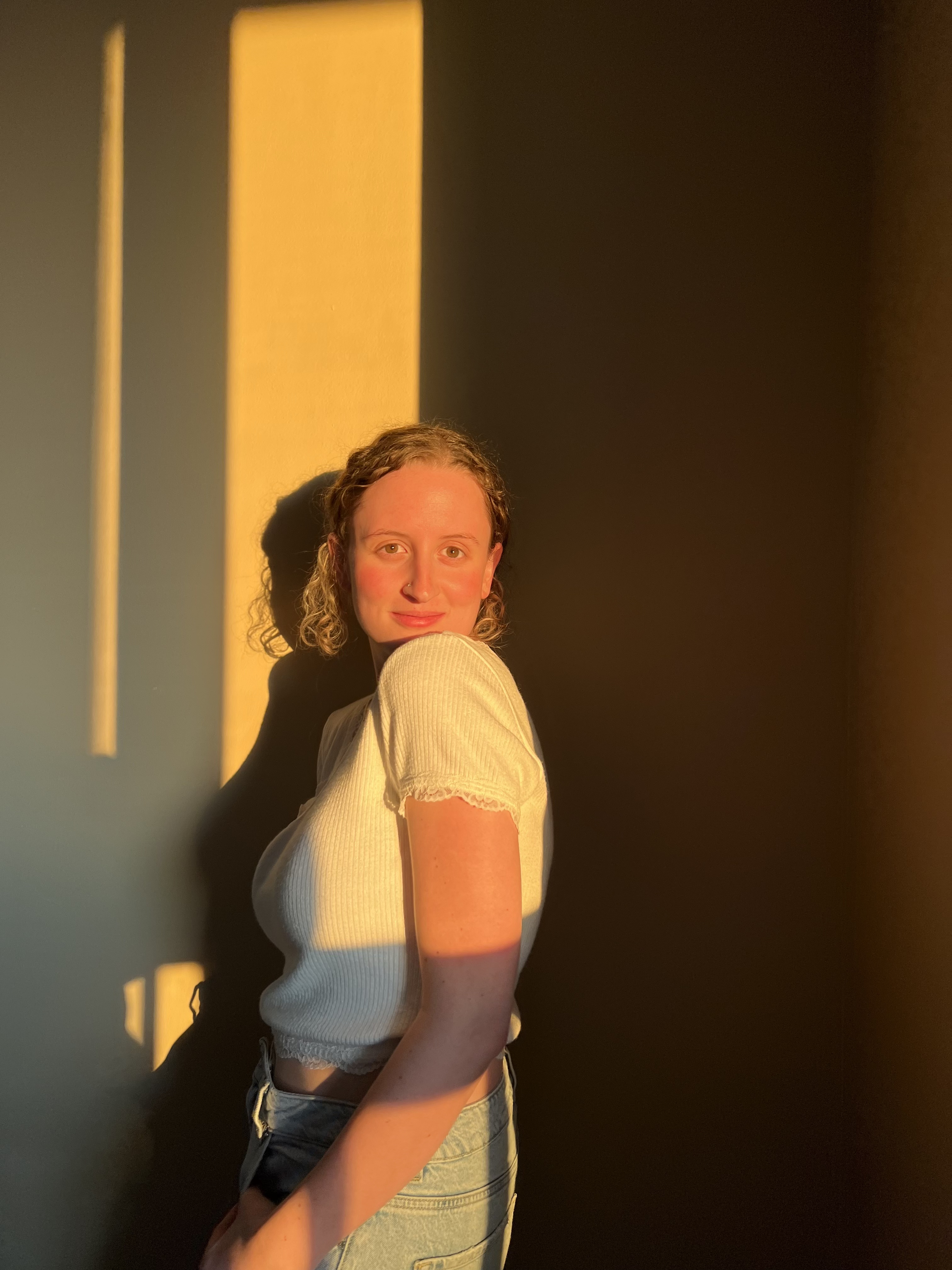 Taylor Gaudon pose en regardant par-dessus son épaule gauche. Le coucher de soleil se reflète sur elle et sur le mur derrière elle.