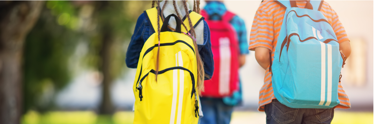 : The backs of three youth walking to school. They are wearing backpacks and are outdoors.