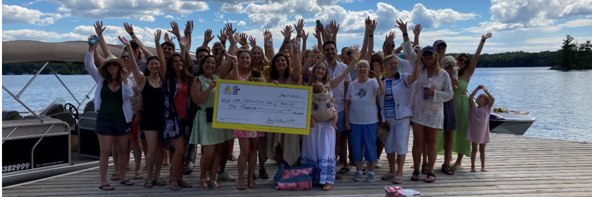 A group of guests stand on top of a waterfront dock and enthusiastically wave at the camera, showcasing their giant-cheque donation of $1,000 