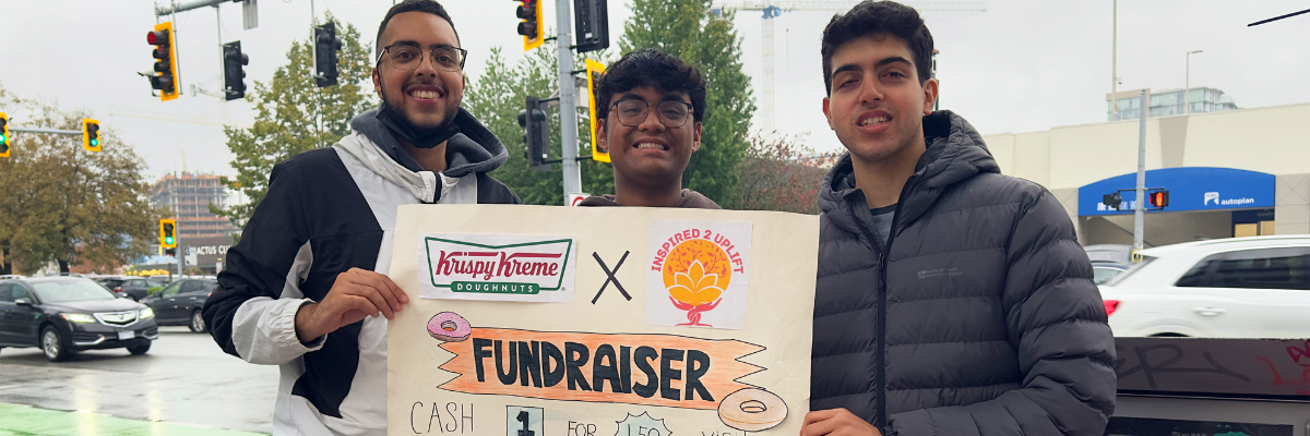 Aidan et ses amis recueillent des fonds pour Inspired 2 Uplift. Ils se tiennent à l’extérieur, devant une table remplie de beignes. Le trio tient une pancarte sur laquelle on peut lire « Krispy Kreme Donuts – Inspired 2 Uplift Fundraiser ».]