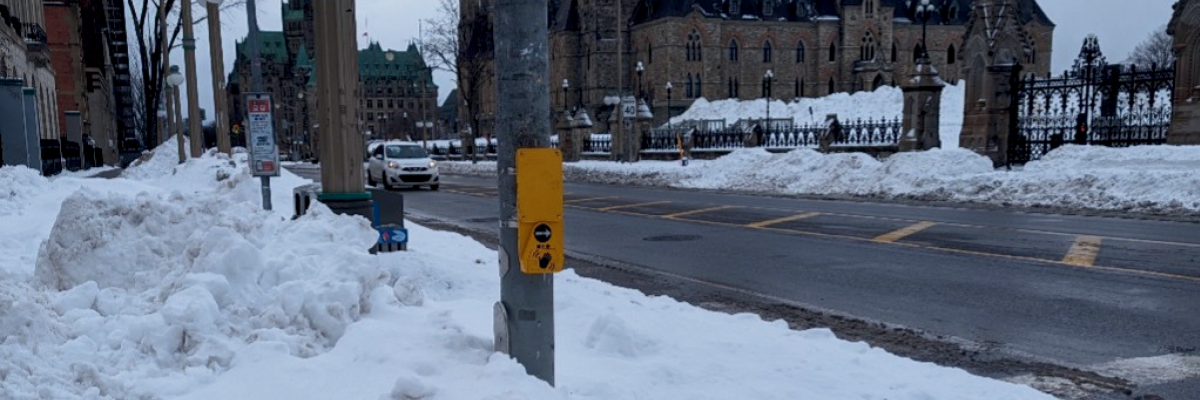 Un bouton de signalisation accessible fixé à un poteau situé sur un passage piéton à Ottawa, avec les bâtiments du Parlement en arrière-plan. Le poteau est entouré d'un gros banc de neige qui atteint presque la hauteur du bouton et s'étend sur environ un mètre vers l'extérieur, ce qui empêche les piétons de l'atteindre.