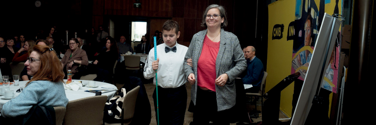 Dawn Pickering smiles as she and her son, Ollie, walk to the stage for the family panel at the Parliamentary launch of CNIB’s Children’s Charter. Attendees and a large CNIB banner are visible in the background. Ollie wears a white dress shirt with a black bowtie and carries a white cane.