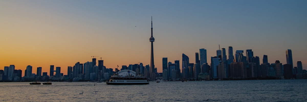 The Toronto skyline at sunset taken from Toronto Island. The sky is a darkening rich blue, and the sunlight in yellow and gold frames the buildings and skyline. A ferry crosses the harbourfront.
