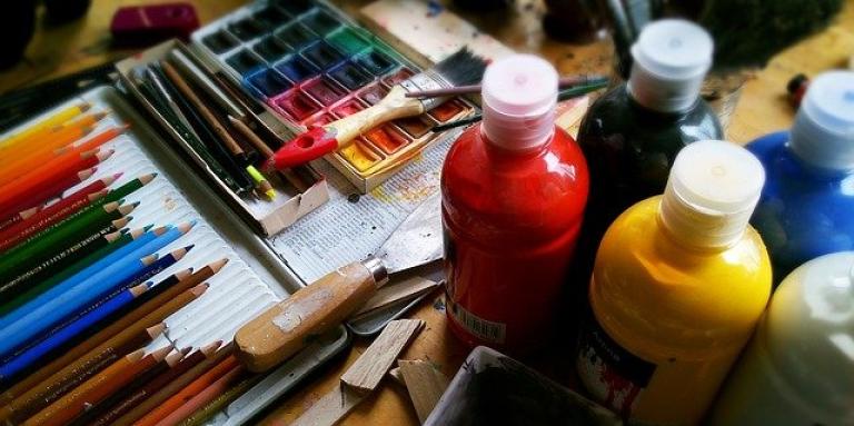 A tabletop covered in art supplies; coloured pencils, paintbrushes and jugs of paint.