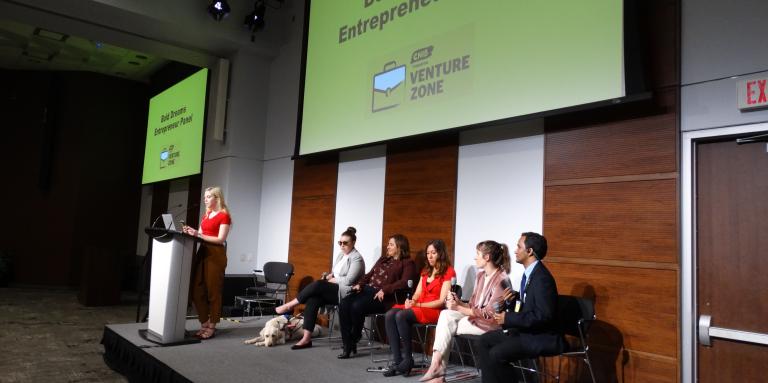 A group of panelists sits on stage at Connecting the Dots. Behind them is a slideshow screen which displays "Bold Dreams - Entrepreneur Panel."