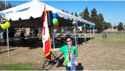 Janna est debout à côté d'un drapeau canadien à l'extérieur d'une grande tente. Elle sourit et tient un pompon dans sa main gauche.