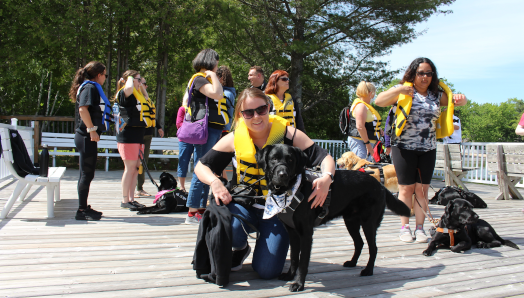  group of guide dog handlers wearing lifejackets stand on the dock at CNIB Lake Joe with their guide dogs. Taylor crouches and poses in the foreground next to her guide dog, Wallace.