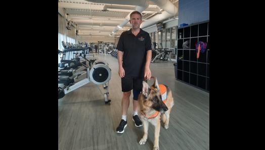 Bob and his German Shepherd guide dog stand inside a gym. The gym is filled with workout equipment.