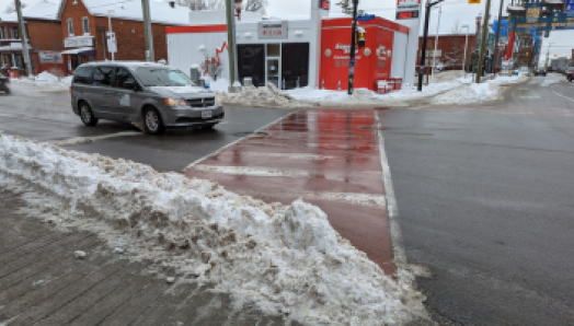 A pile of plowed snow blocks the sidewalk at a busy crosswalk