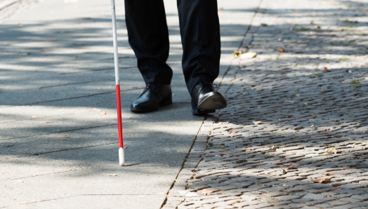 A white cane scans a sidewalk. In the background, men's shoes trail behind the tip of the cane.