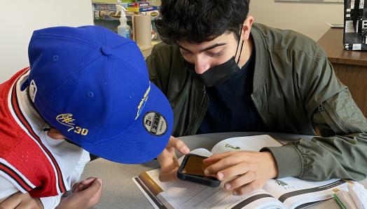 In a classroom, Aidan sits with a young student and shows him the magnification features on his iPhone. Aidan is holding his iPhone over a textbook.  The student intently leans-in for the demonstration.