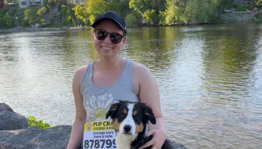 Keely and her border collie puppy sit atop a rock on the shorelines of a lake. Keely is wearing a CNIB pup crawl racing bib and has her arm around the puppy. 