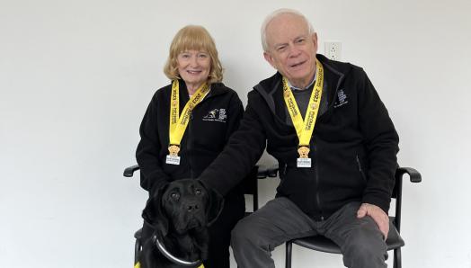 Mary and John Crocker sit to pose wearing 2023 Pup Crawl medals. They are accompanied by a black Labrador Retriever wearing a CNIB Guide Dogs vest.