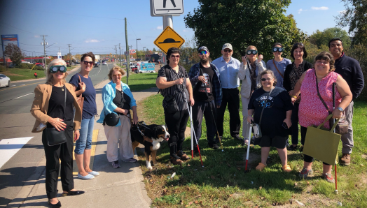 A group of people with sight loss stand next to a pedestrian crossover with public officials in Moncton during the Fall 2023 Get on Board campaign.