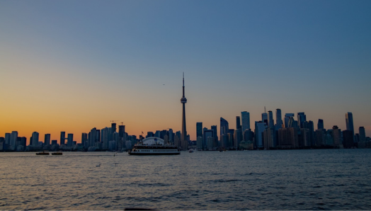 La ligne d'horizon de Toronto au coucher du soleil, prise depuis l'île de Toronto. Le ciel est bleu foncé, et la lumière du soleil, jaune et or, encadre les édifices et la ligne d'horizon. Un traversier avance dans le Port.