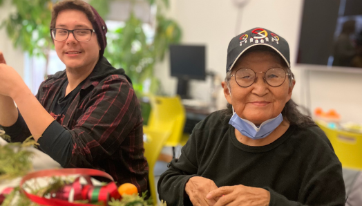 Two Indigenous participants sit at a table in CNIB Thunder Bay smiling. During a holiday program, they create decorations out of fresh cedar bows, dried orange slices, homemade gingerbread cookies, ribbons, bells, cinnamon sticks, and jute twine.