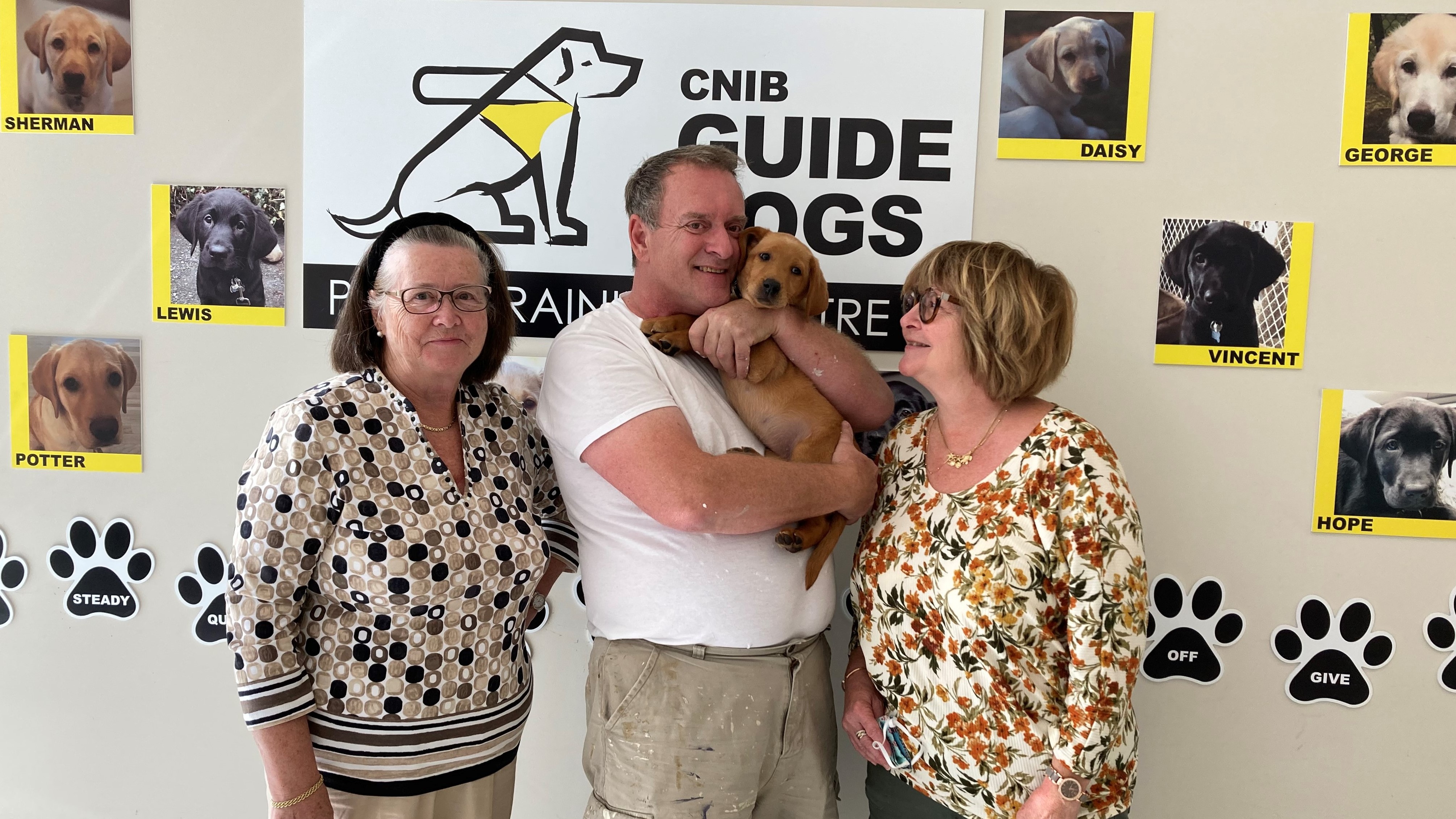Siblings Donna, Ian, and Heather MacKenzie stand together in front of the CNIB Guide Dogs wall in the Halifax office. Ian holds future guide dog Dorrie in his arms.