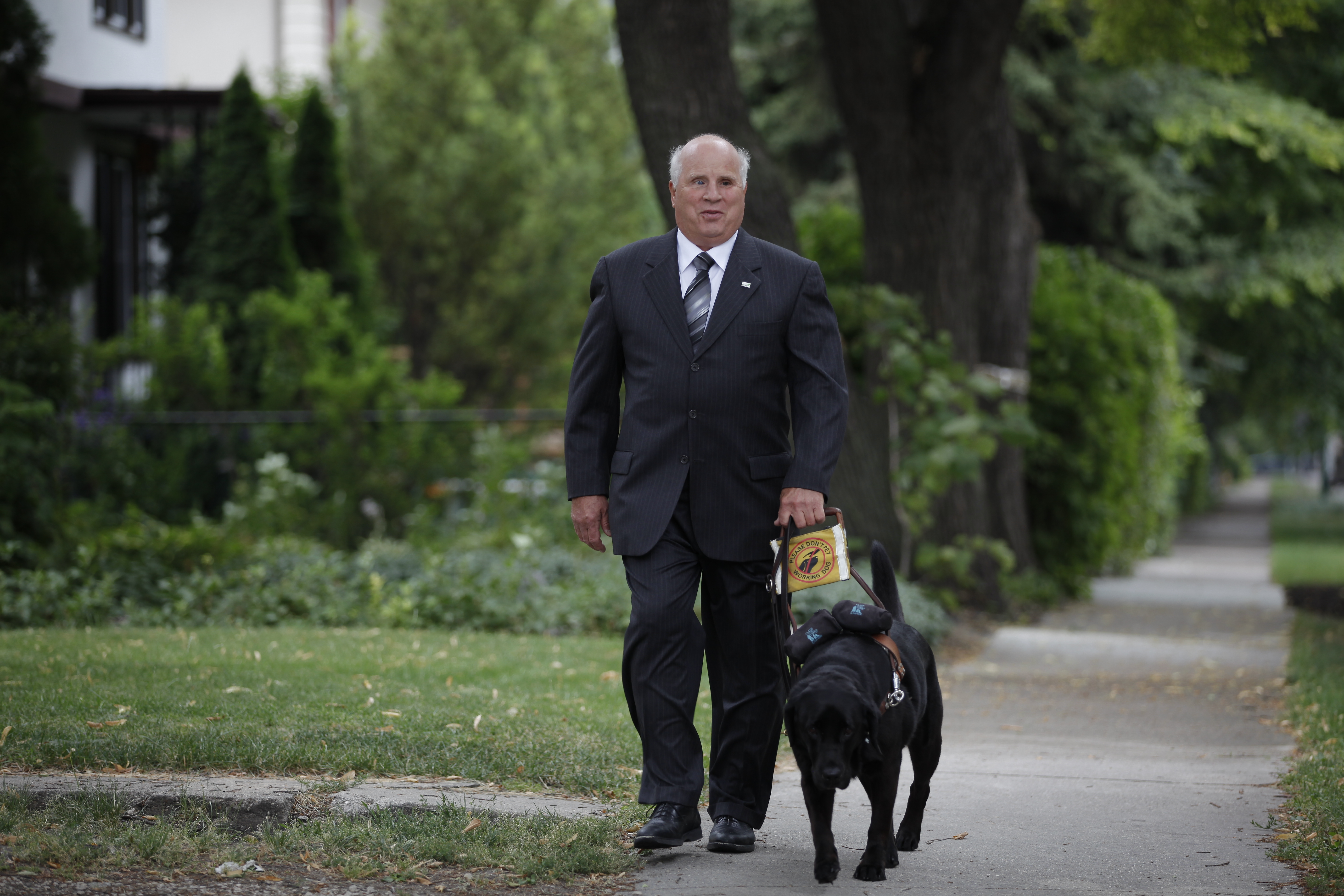 Shep Shell, wearing a black suit, while walking with his guide dog, a Black Labrador.