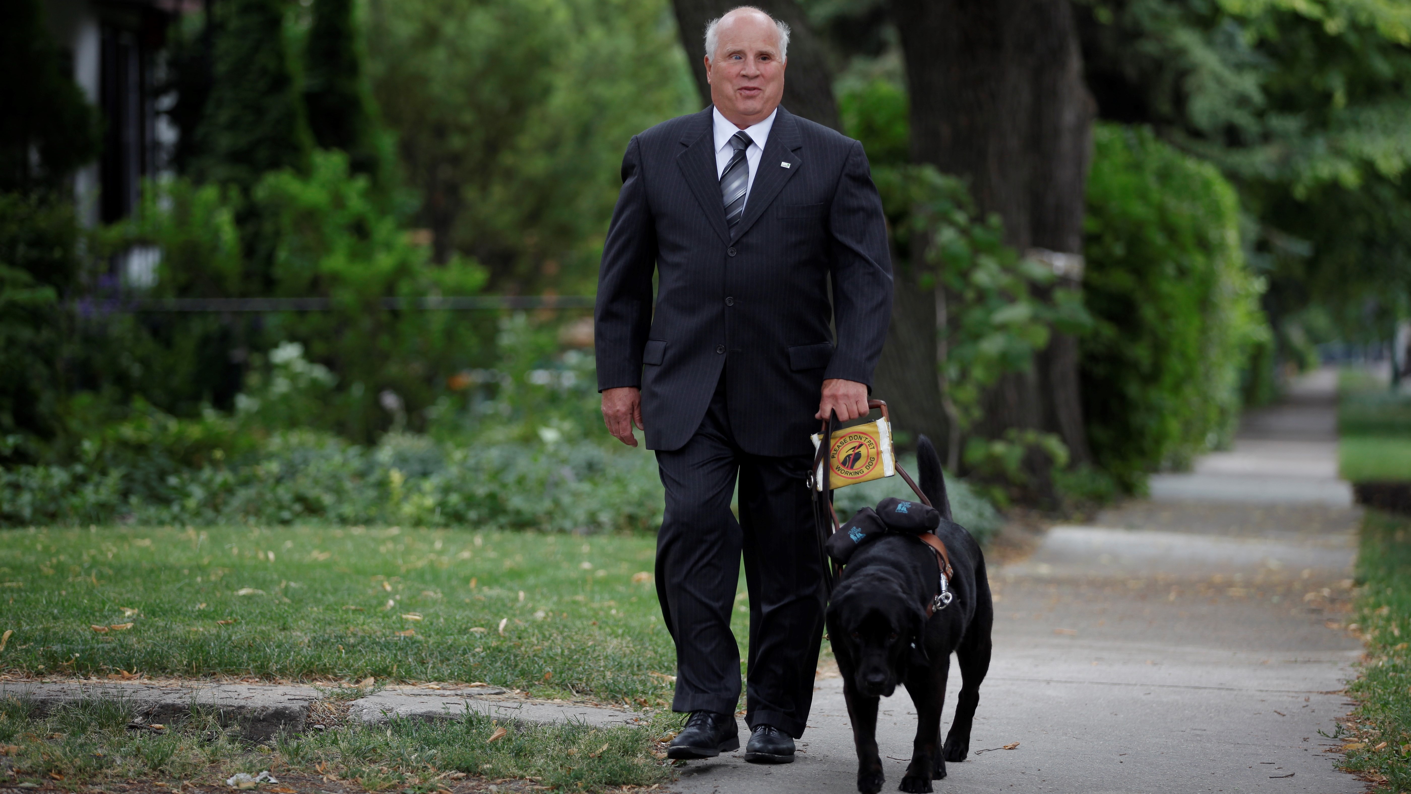 Shep Shell walks with his guide dog whilst wearing a dark suit and tie.