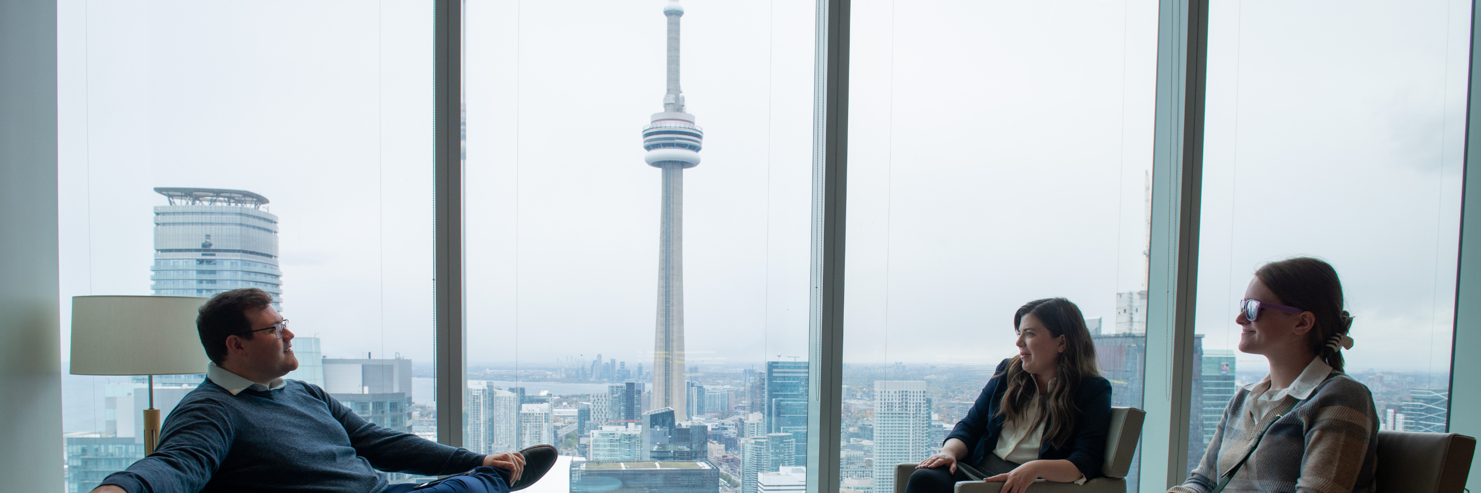 three people sit in an office with the Toronto skyline behind them. 