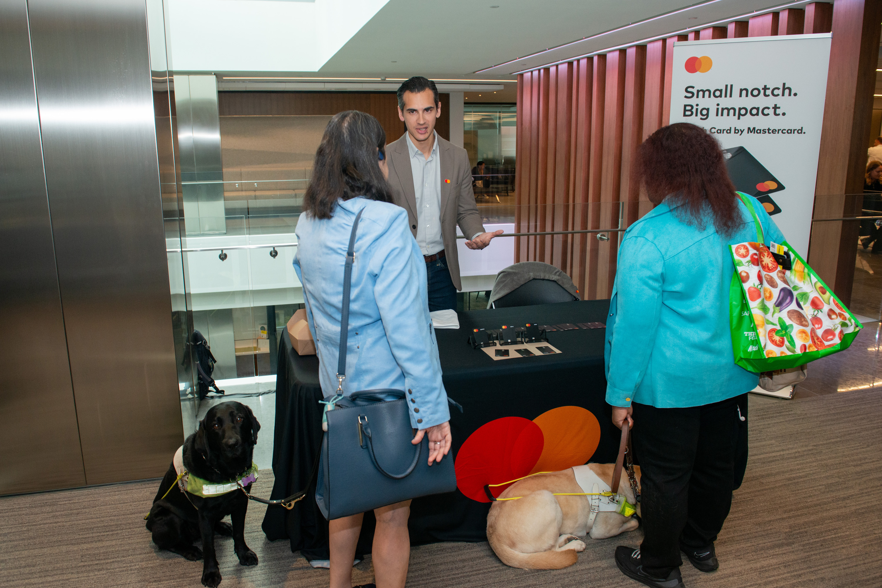 Two people visit a mastercard booth. There is a black lab guide dog sitting beside one of the people visiting the booth. 