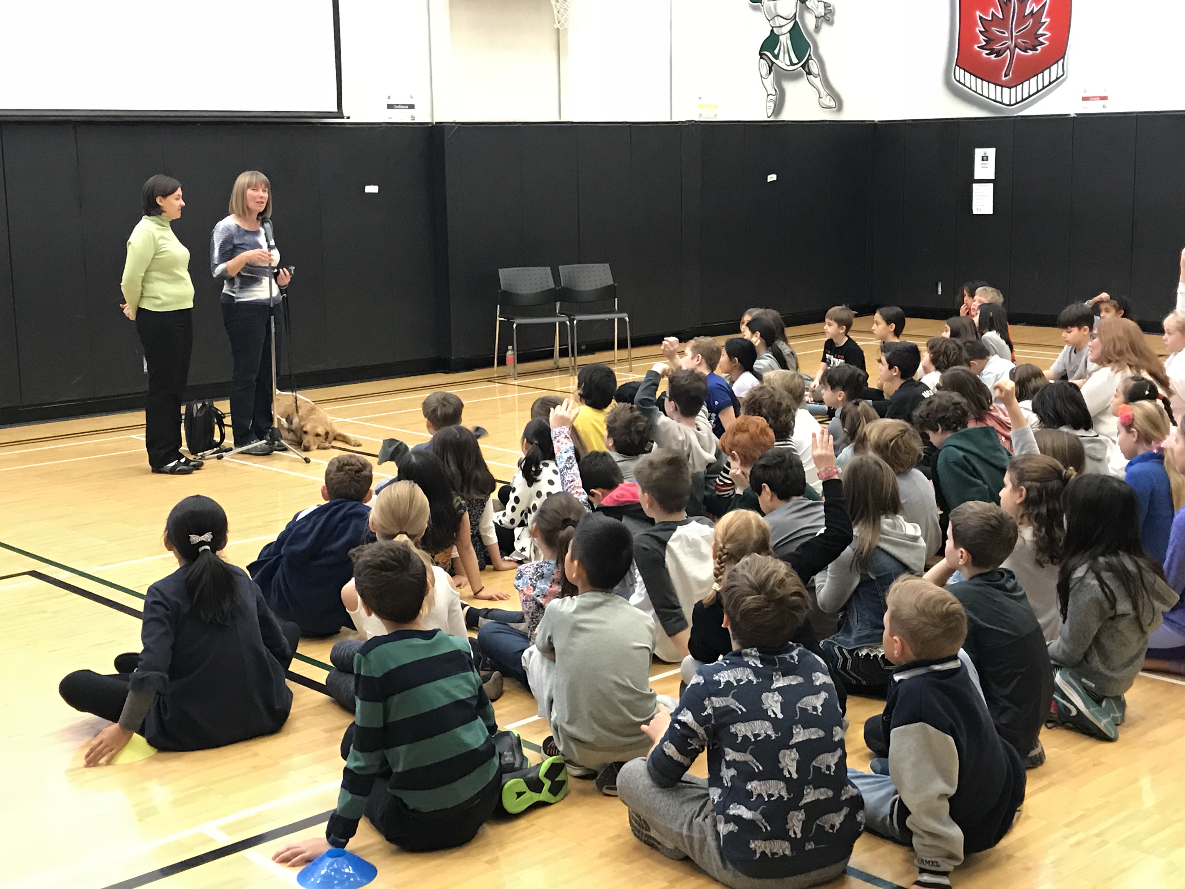 Two Community Engagement Volunteers deliver a presentation in front of a large group of school-aged children sitting in a gymnasium.