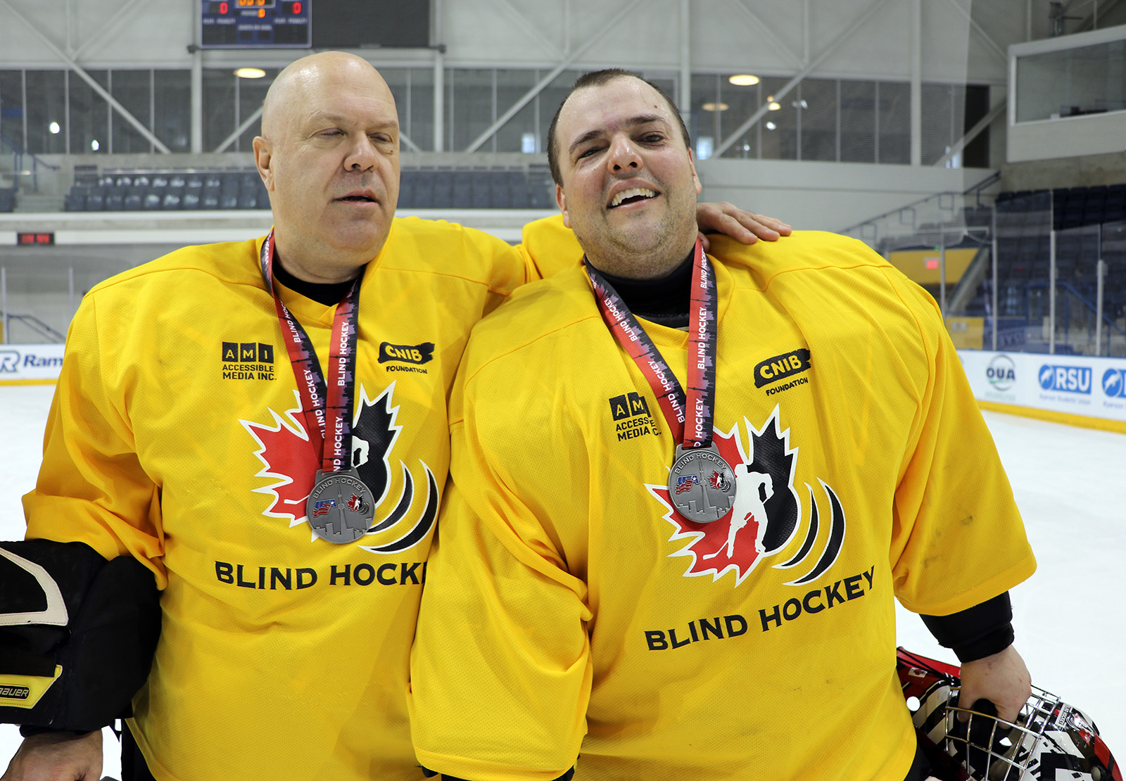 Two adult male Blind Hockey goalies stand on the ice side by side, wearing jerseys, hockey pads, and medals around their necks