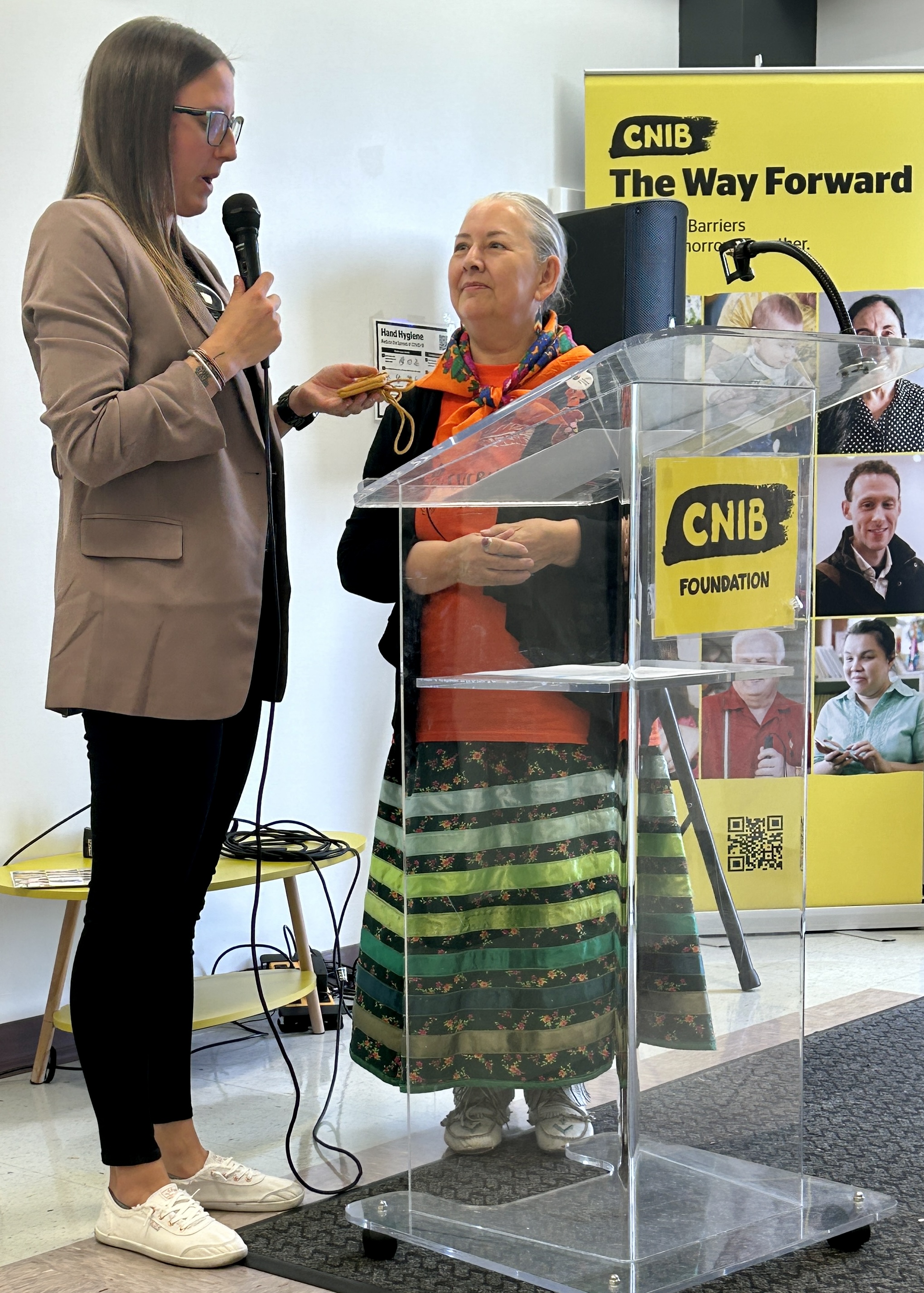A woman wearing a blazer stands next to a female Indigenous elder at a podium with a CNIB banner in the background