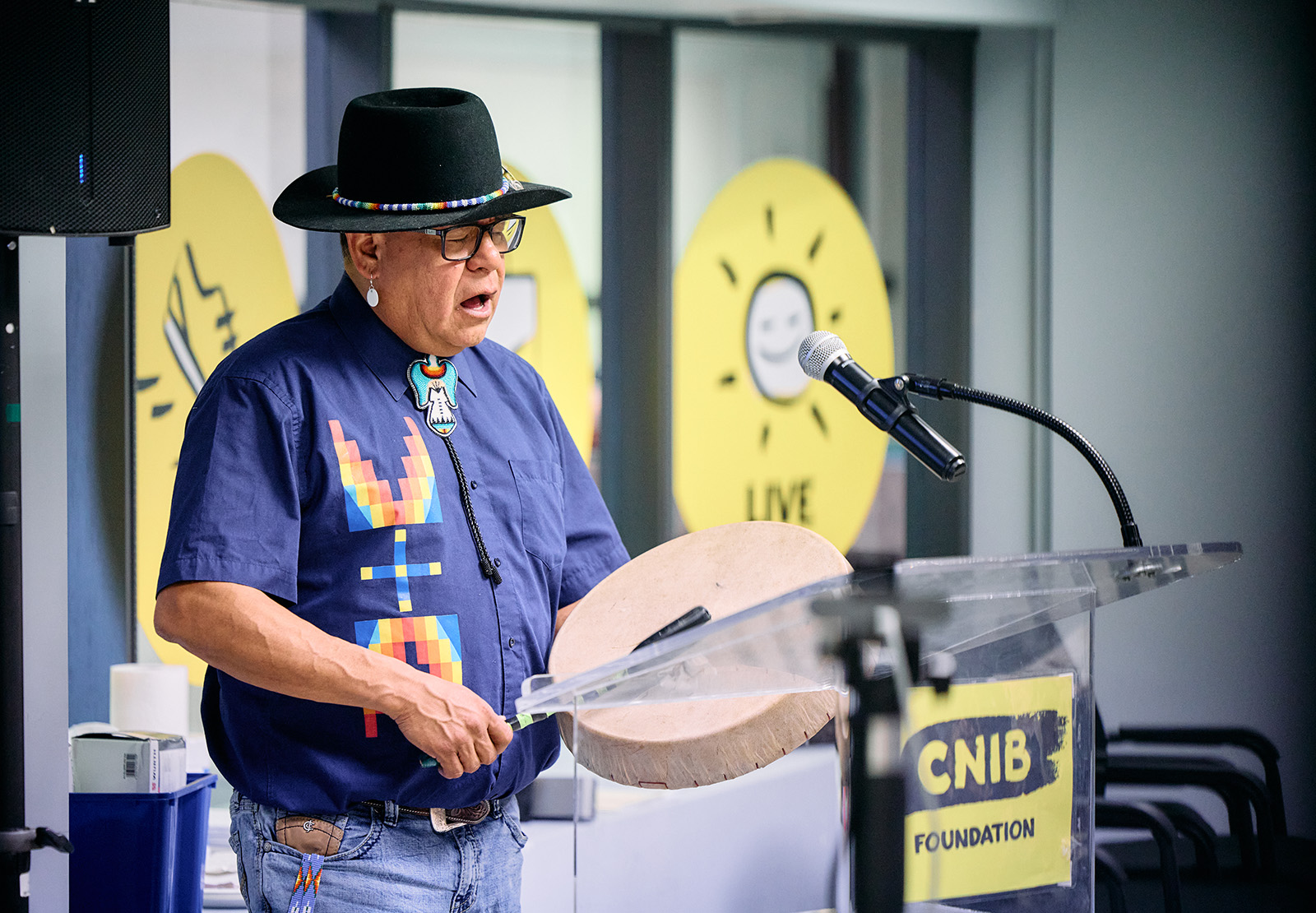 An Indigenous man sings while playing a drum, standing at a podium with the CNIB logo 