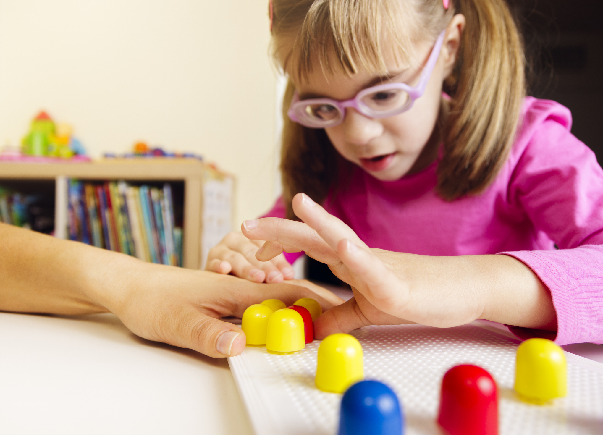 Young girl with pink glasses feels primary coloured pegs while an adult guides her hands