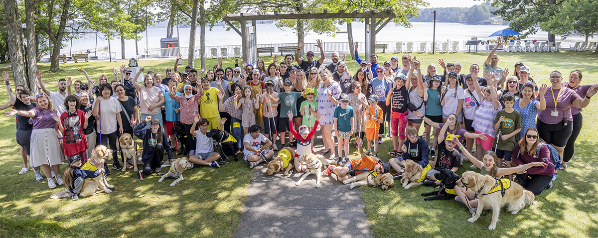 A group shot of campers, staff and buddy dogs all gathered together on the lawn in front of the waterfront. Everyone is smiling and has their hands raised in the air in celebration