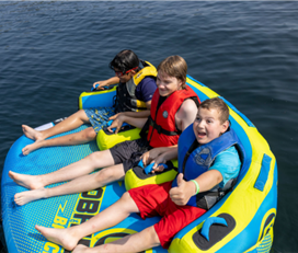 Three young campers sit on an inflatable inner tube on the water. One camper has his thumb up and has a big smile on his face.