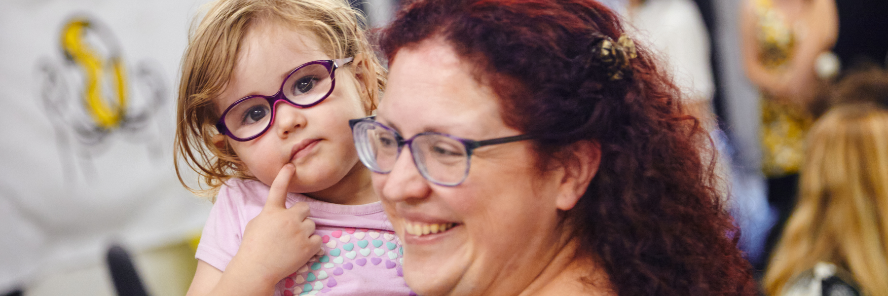 A little girl with glasses points to her chin, while her mother holds her in her arms.