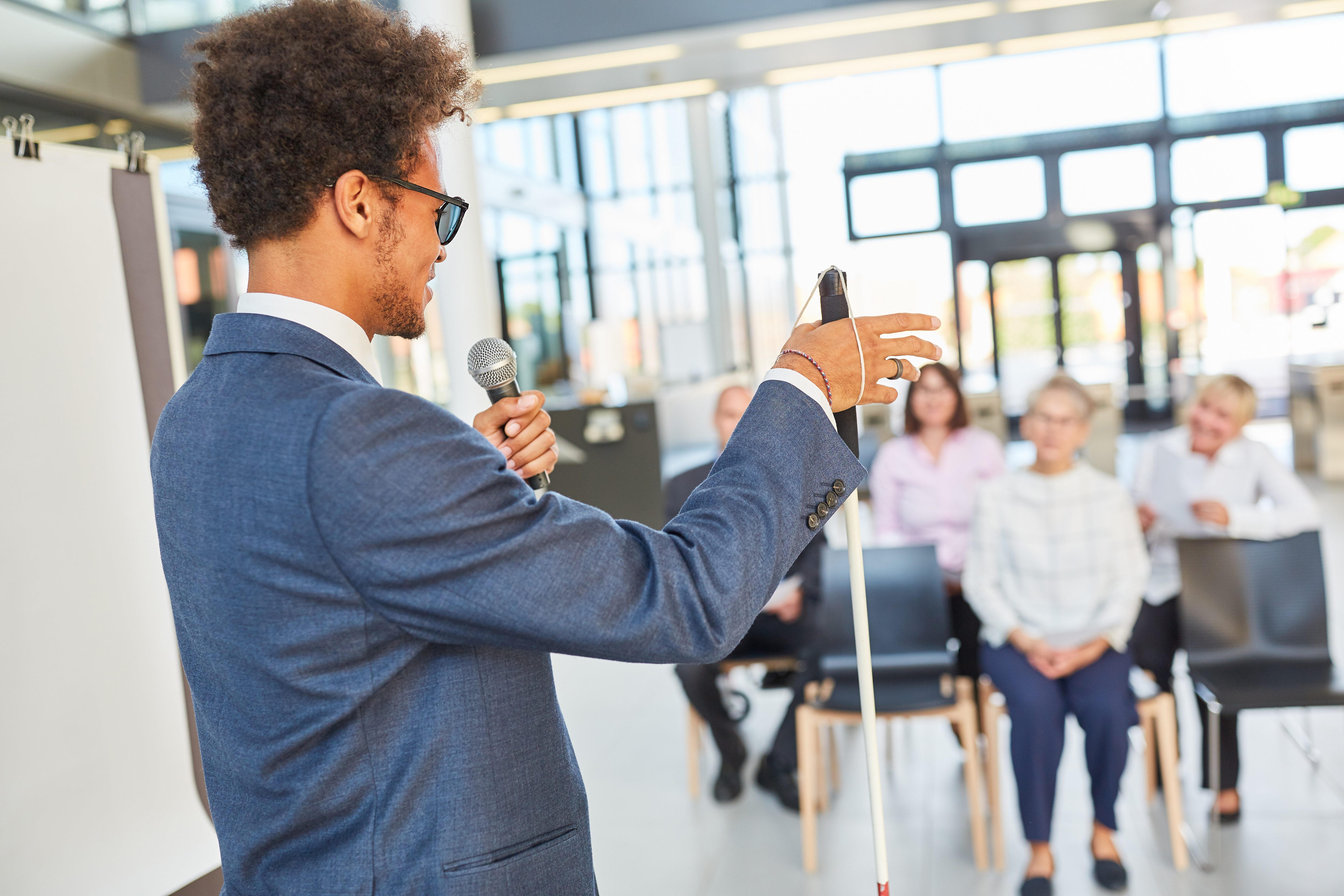 A young man stands at the front of a meeting room and speaks to a group of people. He is holding a microphone in his left hand and a white cane in his right hand.