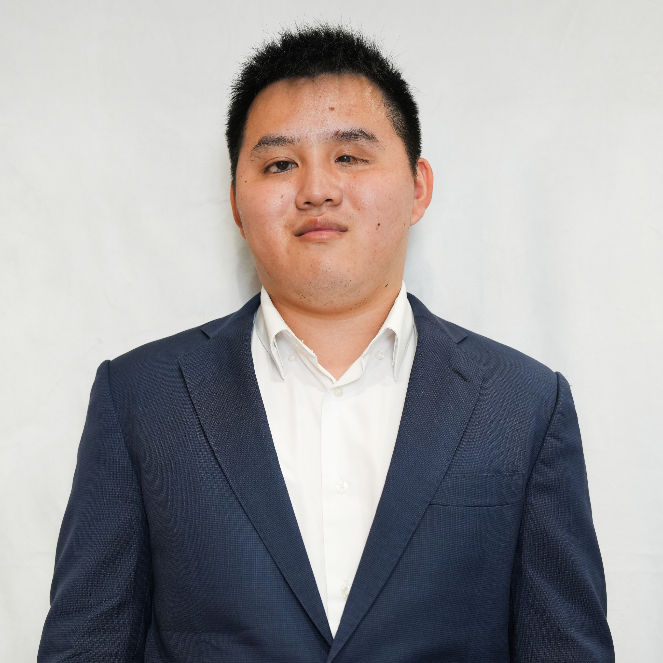 Zhicheng, a young Asian man wearing a navy suit with a white dress shirt, stands in-front of a white background. Zhicheng is smiling and the photograph resembles a professional headshot. 