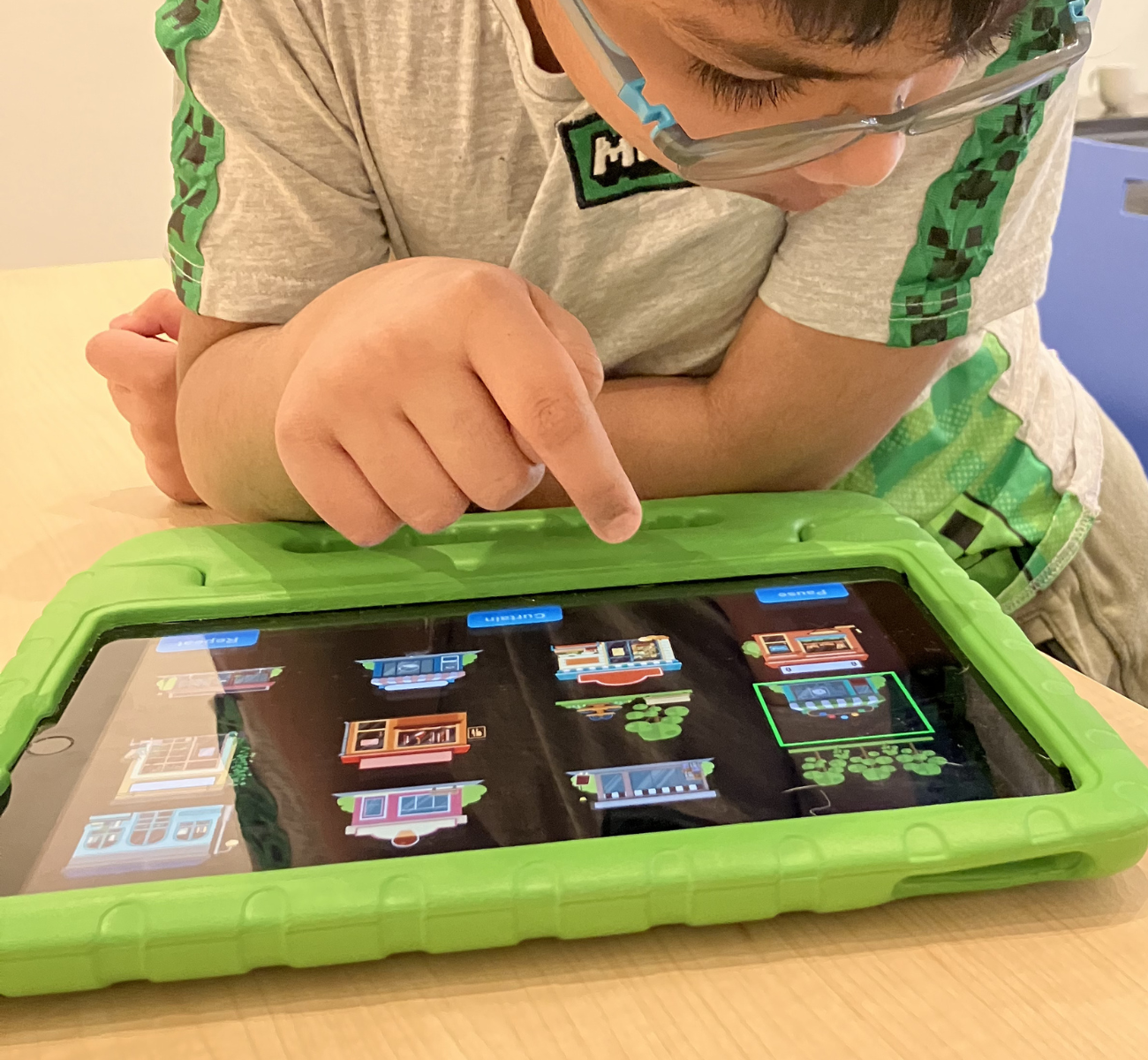 A young boy plays on a tablet. The tablet is sitting on top of a table in a protective green rubber case. The child tapping on the tablet screen and interacting with the FirstTap app.