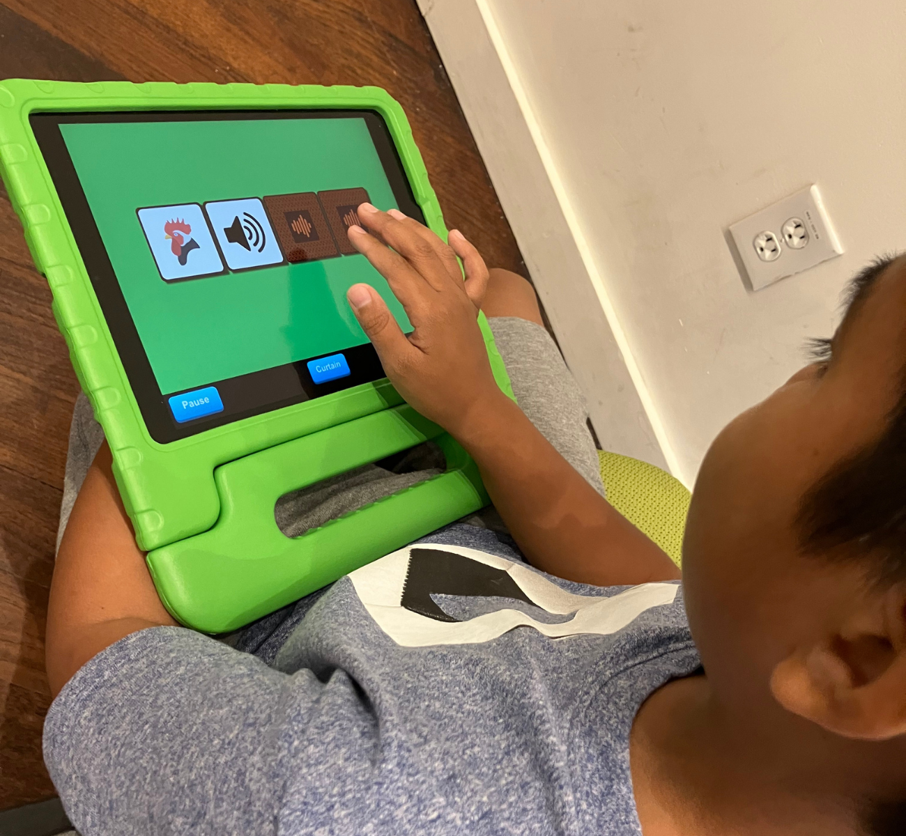 A young boy plays on a tablet. He is sitting on the ground, and the tablet is in his lap is in a protective green rubber case. The picture is an overhead view of the child tapping on the tablet screen and interacting with the FirstTap app