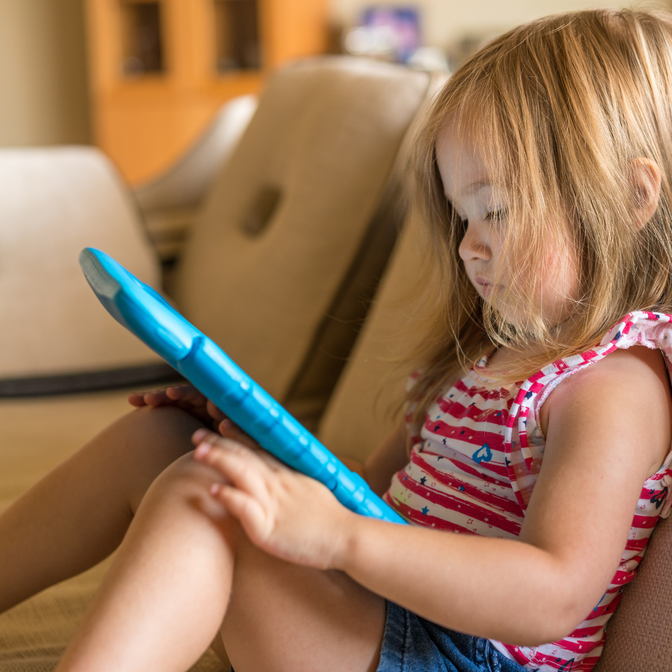 A young girl sits on a couch and plays on a tablet. The tablet is in a protective blue rubber case.