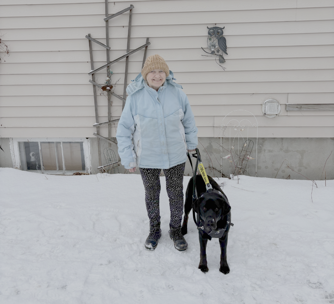 Barb stands in a snowy backyard with her black guide dog, Beacon, in harness at her side.