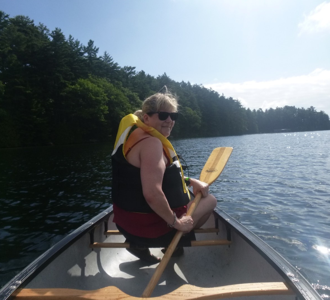 A young woman sits at the front of a canoe. She's holding a paddle and wearing a lifejacket, and turns around to face the camera. The canoe is gliding along the shoreline of Lake Joe.